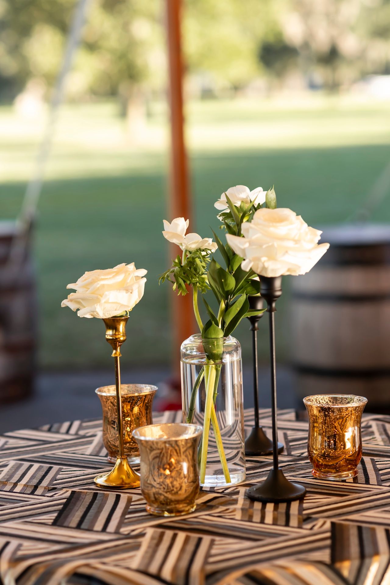 Elegant table centerpiece with peach roses in clear vases and golden mercury glass votives on a geometric patterned tablecloth.