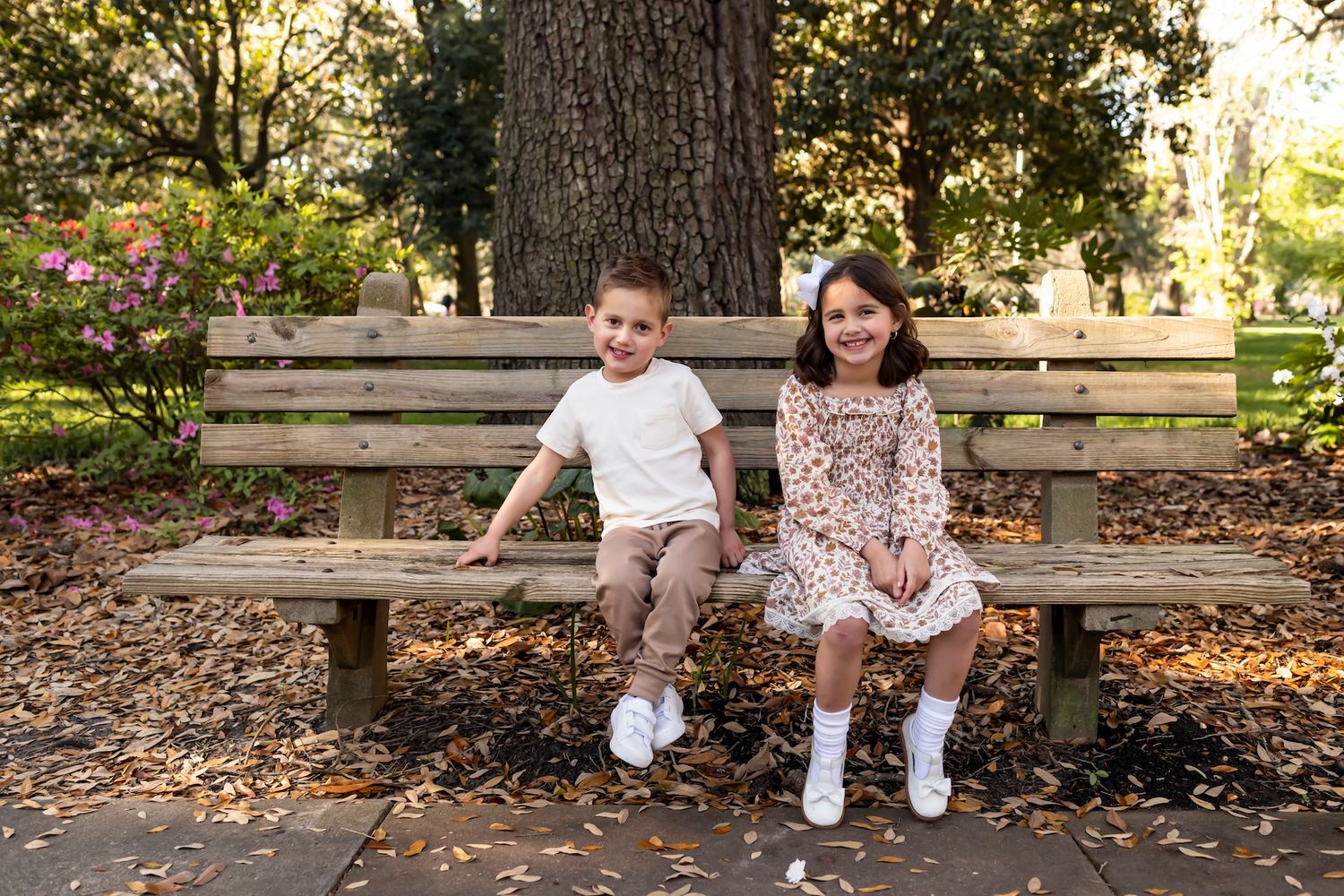 Two children sit together on a wooden park bench surrounded by autumn leaves and trees.