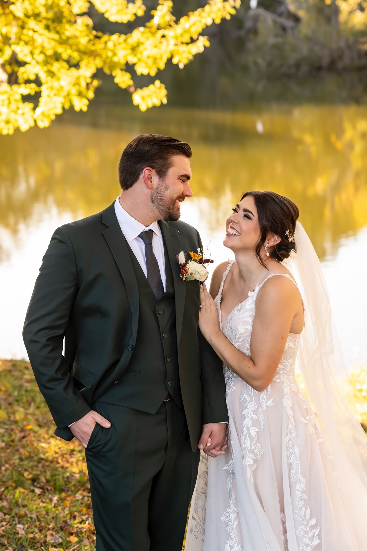 A romantic autumn wedding photo of newlyweds sharing a joyful moment by a golden-lit pond under yellow fall foliage.
