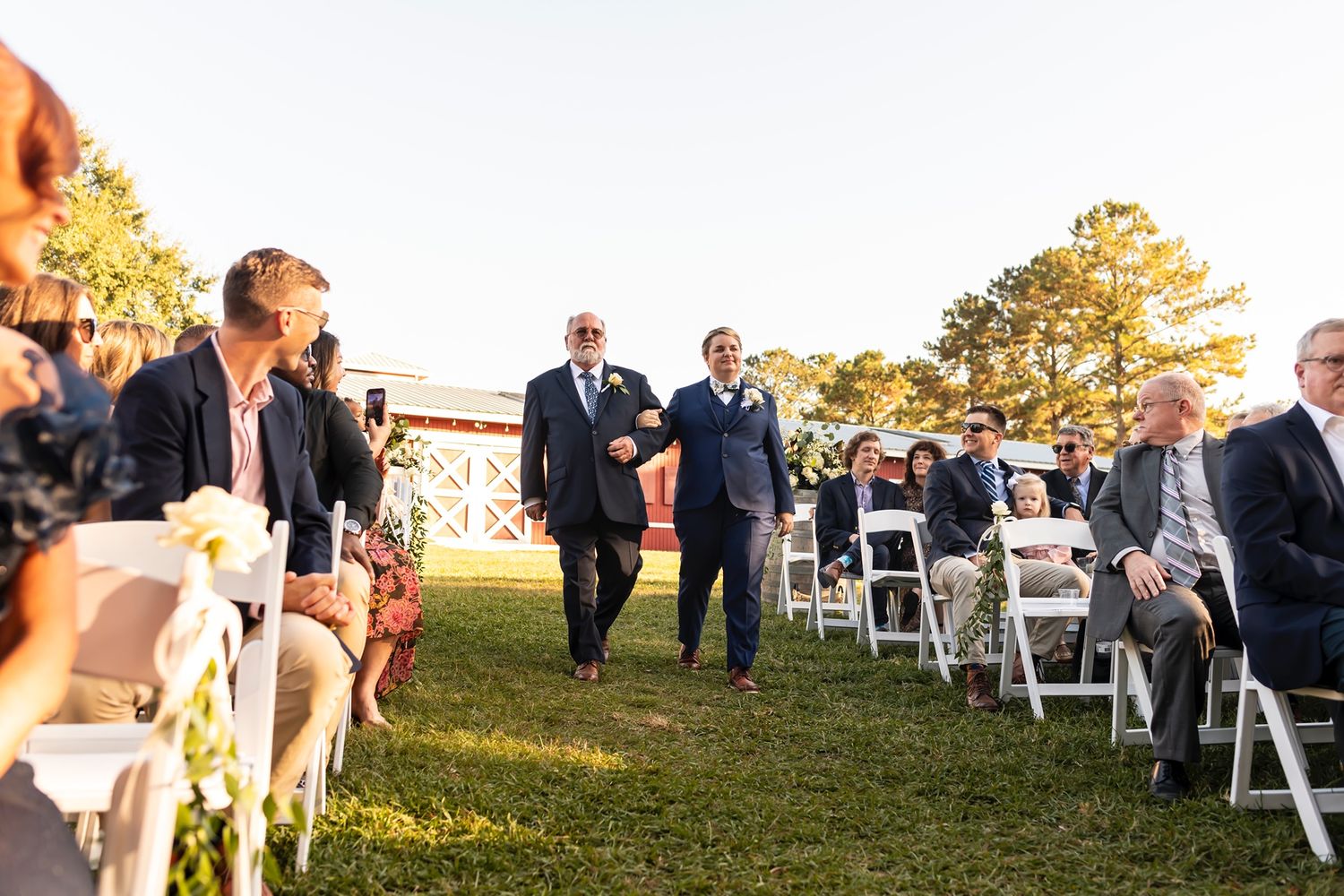 People walking down an outdoor wedding ceremony aisle lined with white chairs on a grassy lawn at sunset.