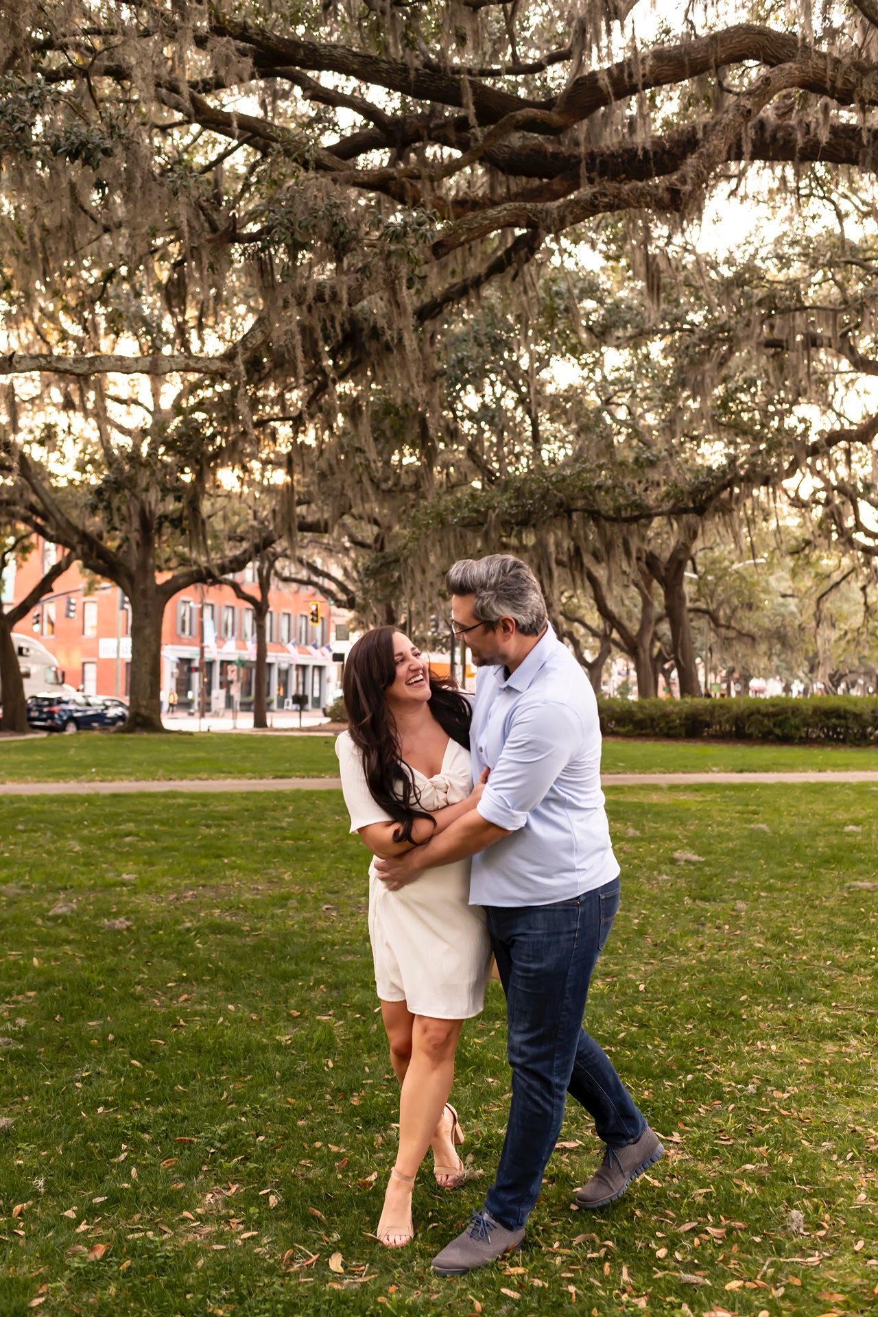 A couple dances romantically beneath Spanish moss draped oak trees on a green lawn with historic buildings in background.