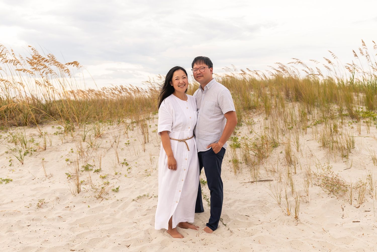 Two people standing together on sandy beach dunes with sea grass swaying in the breeze at sunset.