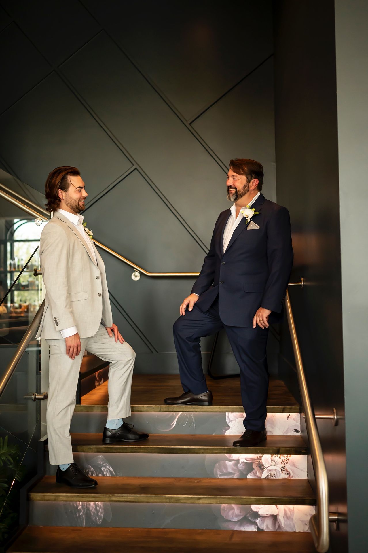 Two people in contrasting suits, one light and one dark, stand on wooden stairs with decorative wall paneling behind them.