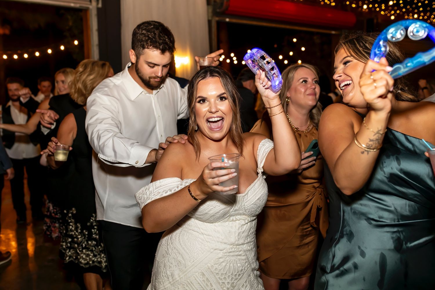 Party guests dance enthusiastically at a wedding reception with festive string lights in the background.