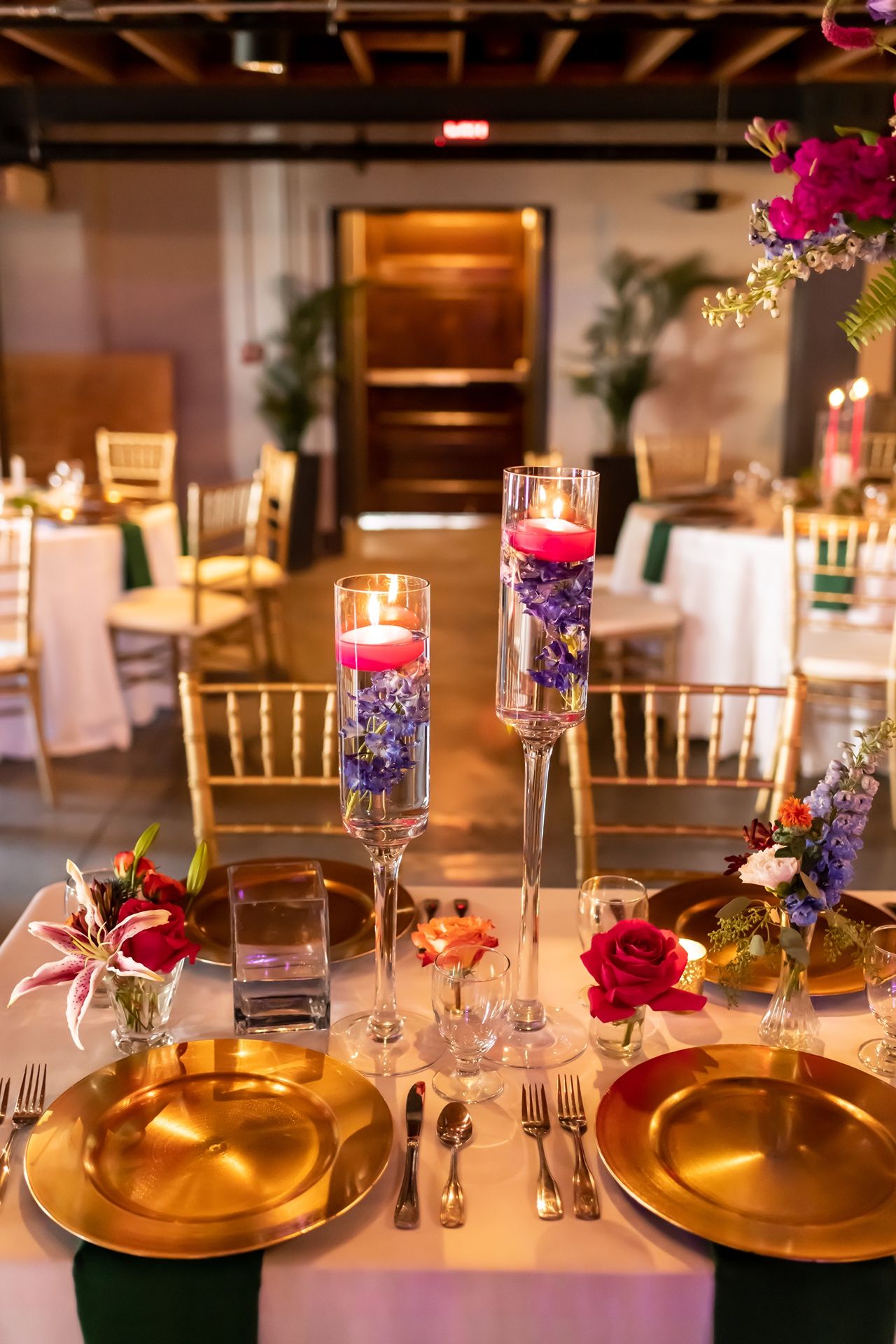 Elegant wedding table setting with gold charger plates, red roses, and purple flowers illuminated by glowing candles.