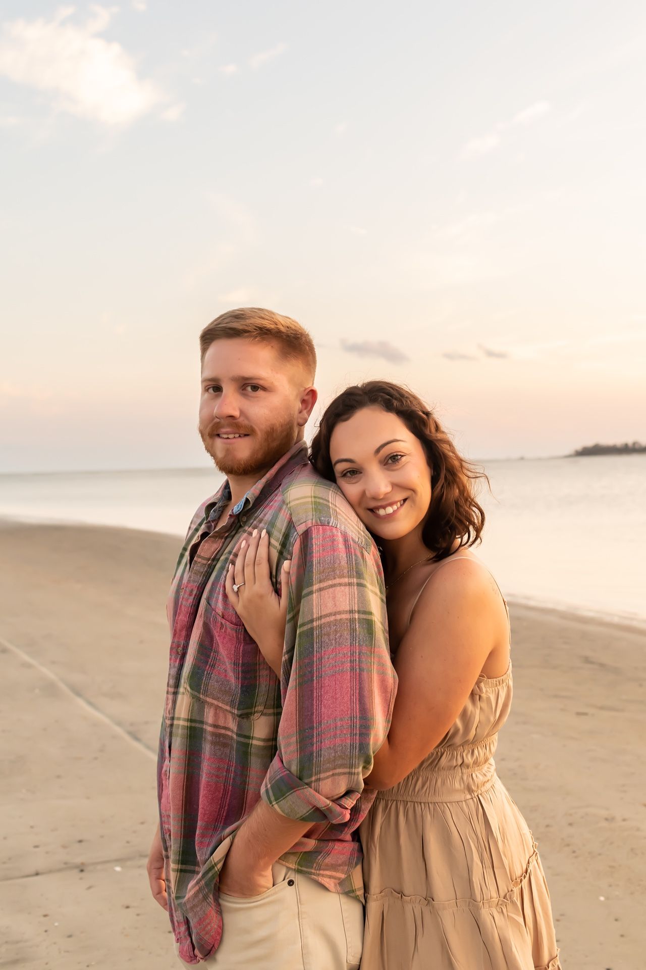Happy couple embracing on a scenic beach at sunset with soft golden lighting and tranquil ocean backdrop.