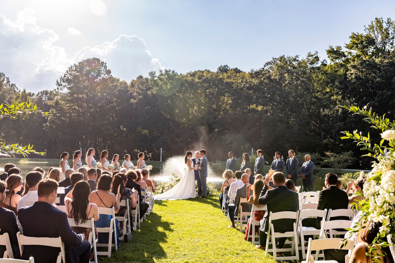 Outdoor wedding ceremony on a sunny lawn bordered by trees with morning mist and guests seated in white chairs.