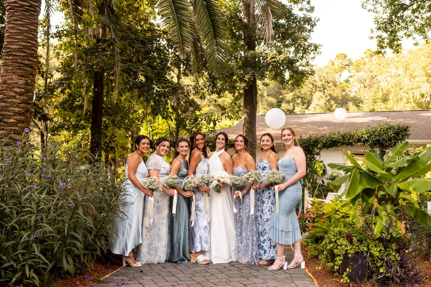 Wedding party in light blue bridesmaid dresses poses together on garden path surrounded by lush greenery and palm trees.