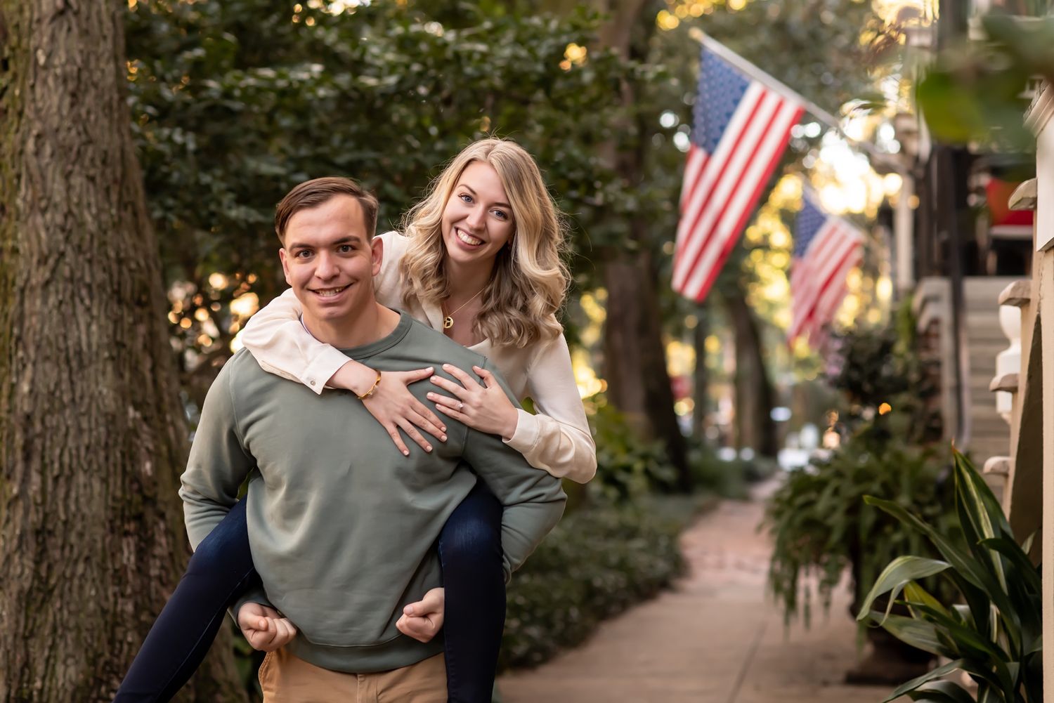 A joyful couple shares a piggyback ride moment on a tree-lined sidewalk with American flags in the background.