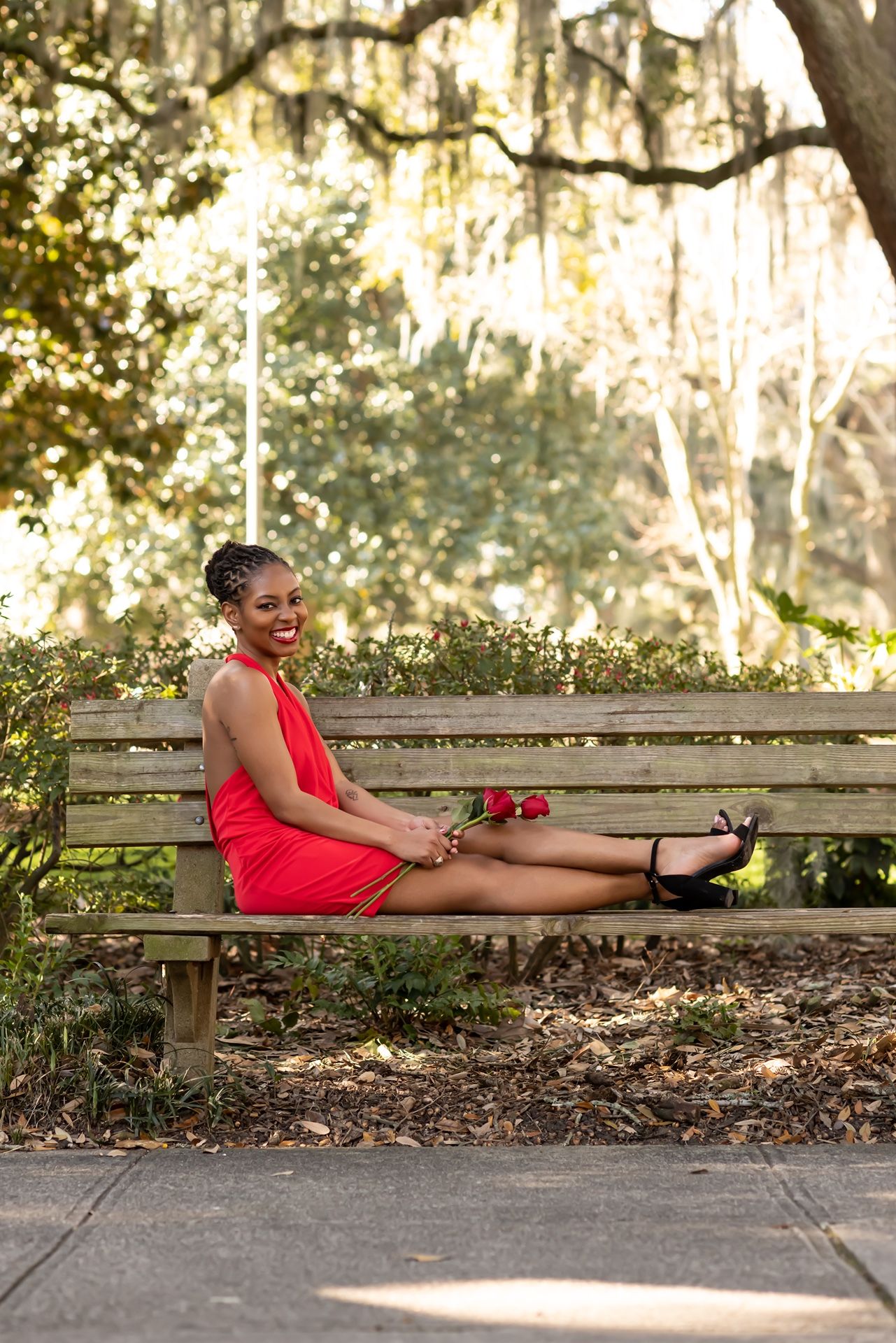Woman in a bright red dress relaxing on a wooden park bench surrounded by Spanish moss and trees.
