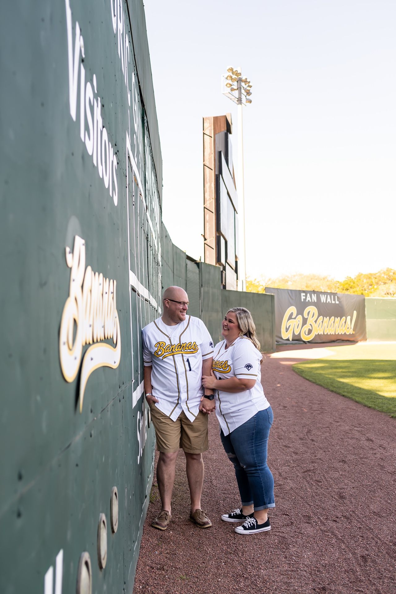 People standing by a baseball field wall, silhouetted against a sunset-lit sports stadium background.