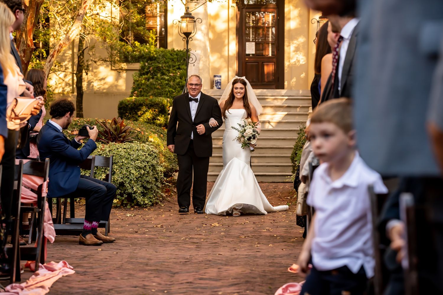 A flower girl walks down an outdoor path while releasing rose petals from a white basket during a wedding ceremony.