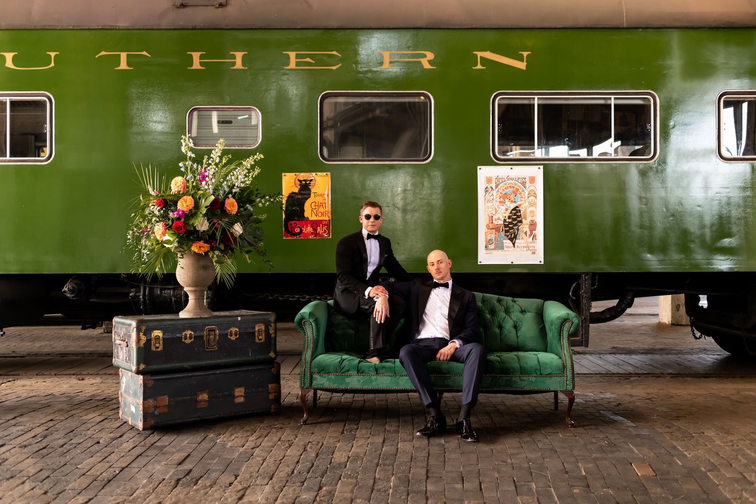 Two grooms sit on a couch in front of a rustic train at Georgia State Railroad Museum.