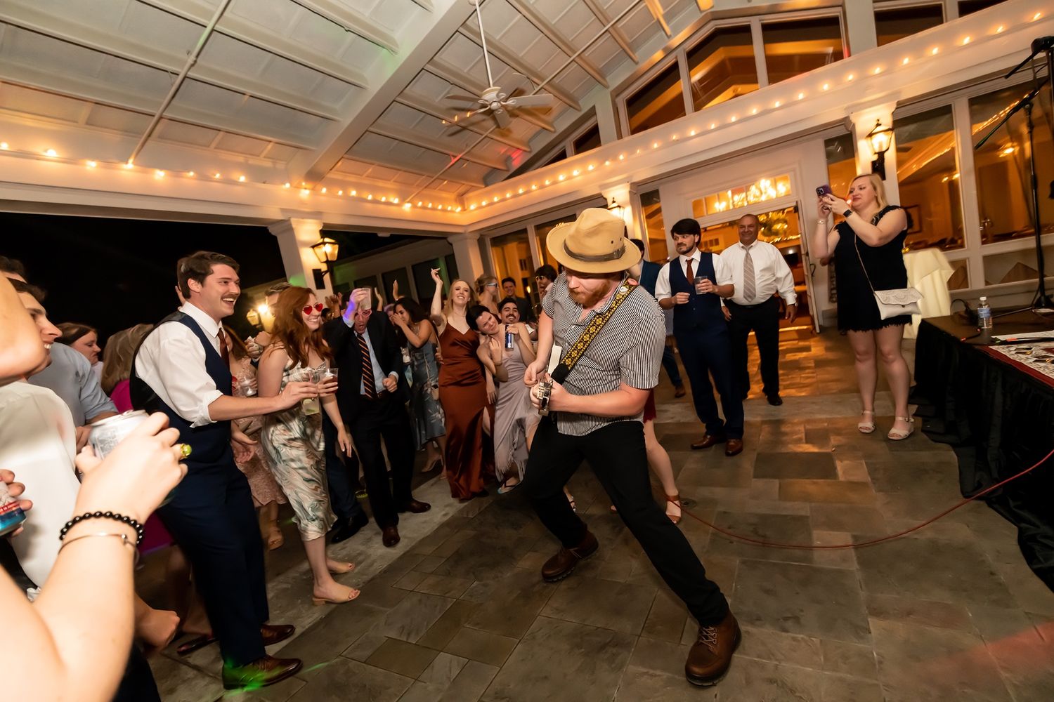 Energetic guests dance on the dance floor during a festive wedding reception in an indoor venue with warm lighting.