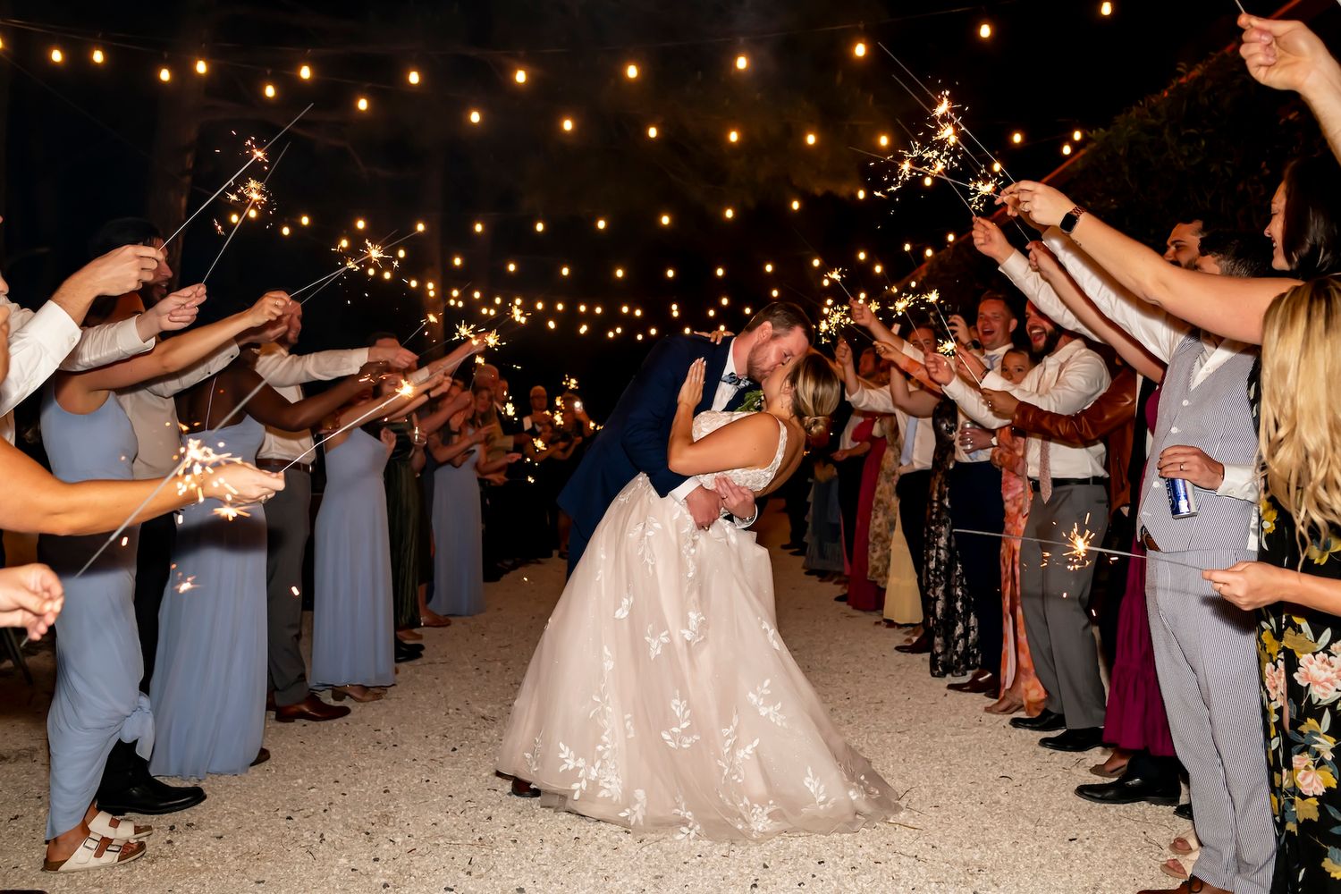 A couple shares a romantic kiss surrounded by guests under twinkling lights at an outdoor evening wedding reception.