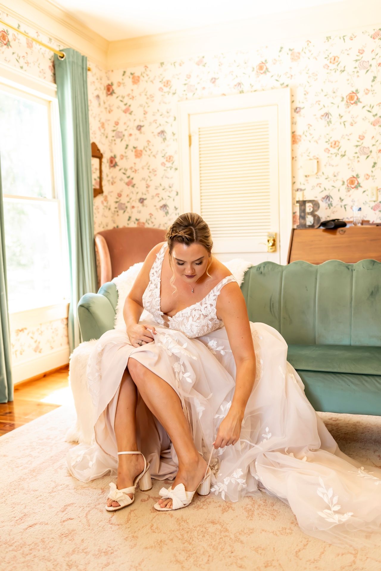 A figure in a white lace wedding dress adjusts a strappy sandal while seated on a velvet sofa in a floral wallpapered room.