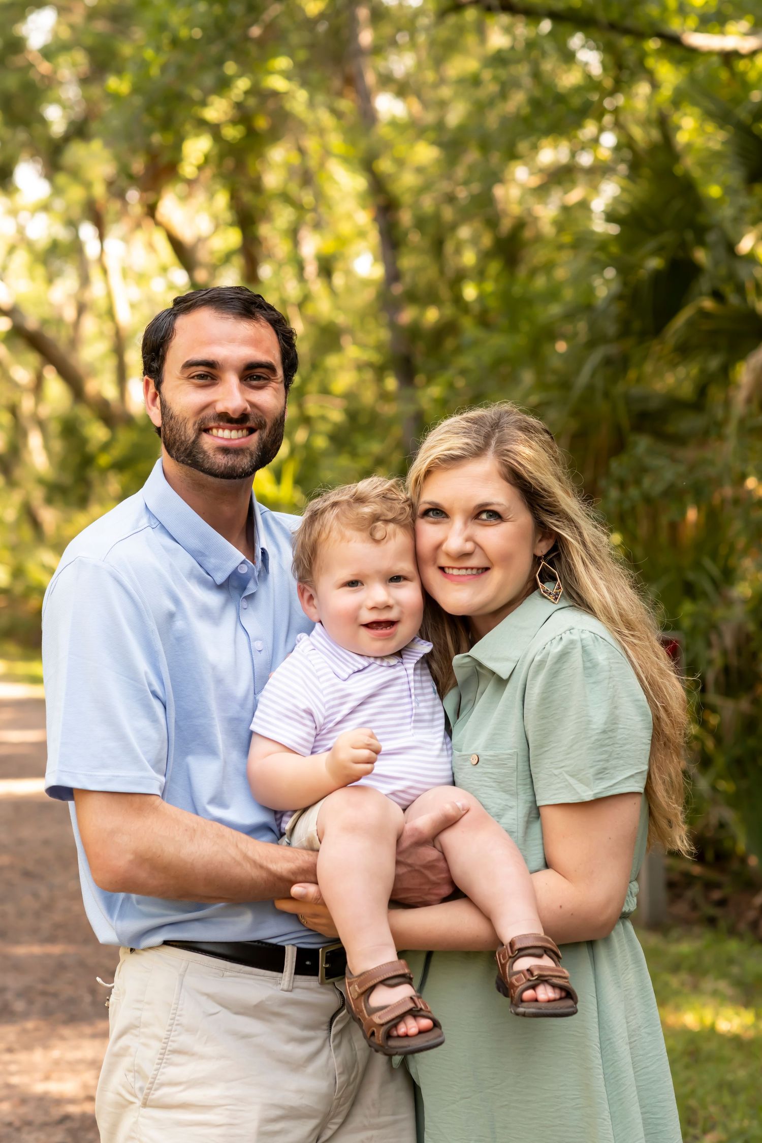 A family poses together outdoors in a wooded setting wearing coordinated casual outfits in blue and sage green tones.