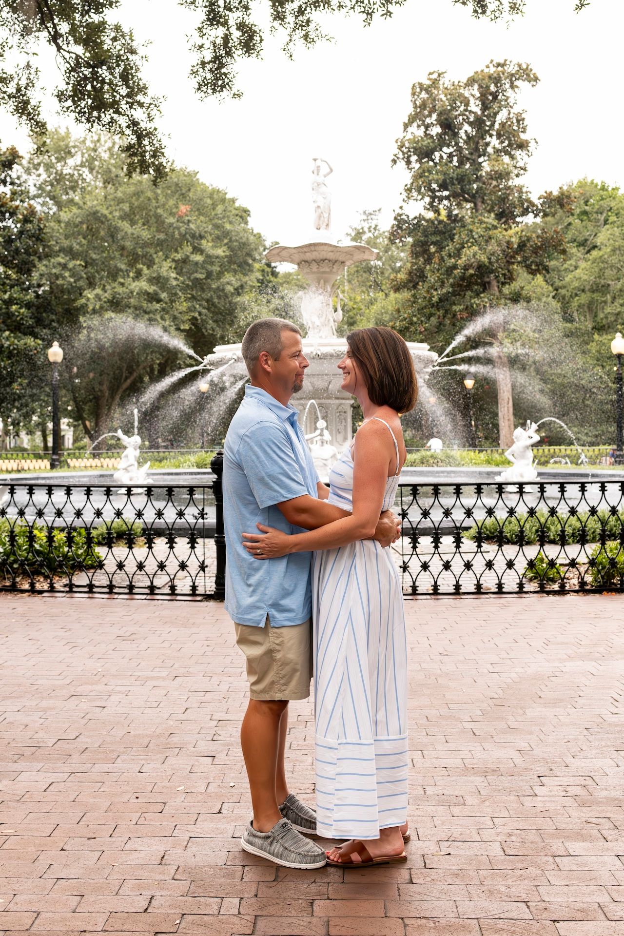 Romantic couple embracing in front of garden fountain with water sprays and iron fencing.