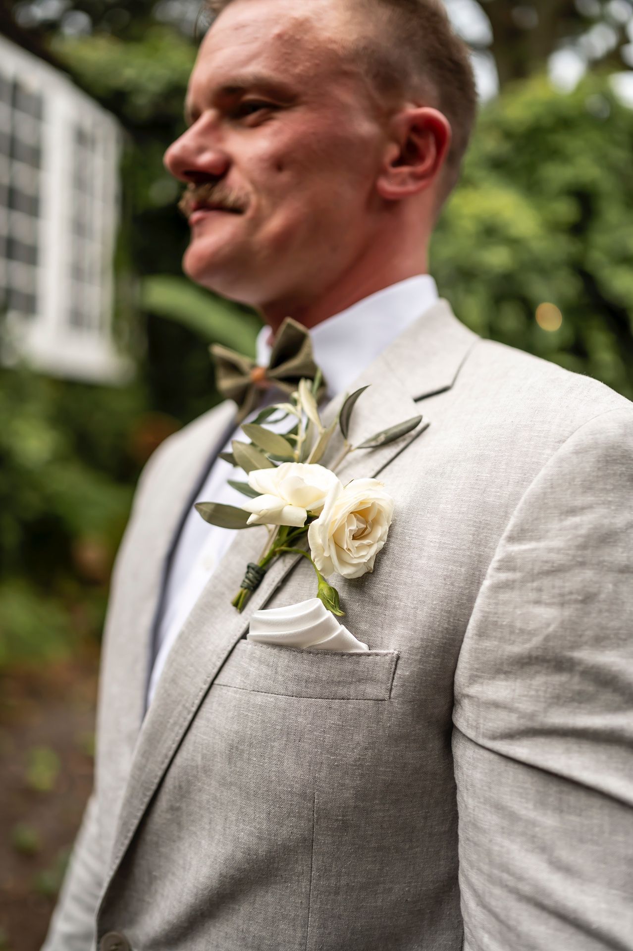 A groom wearing a light gray suit with a white rose boutonniere smiles during an outdoor wedding ceremony.
