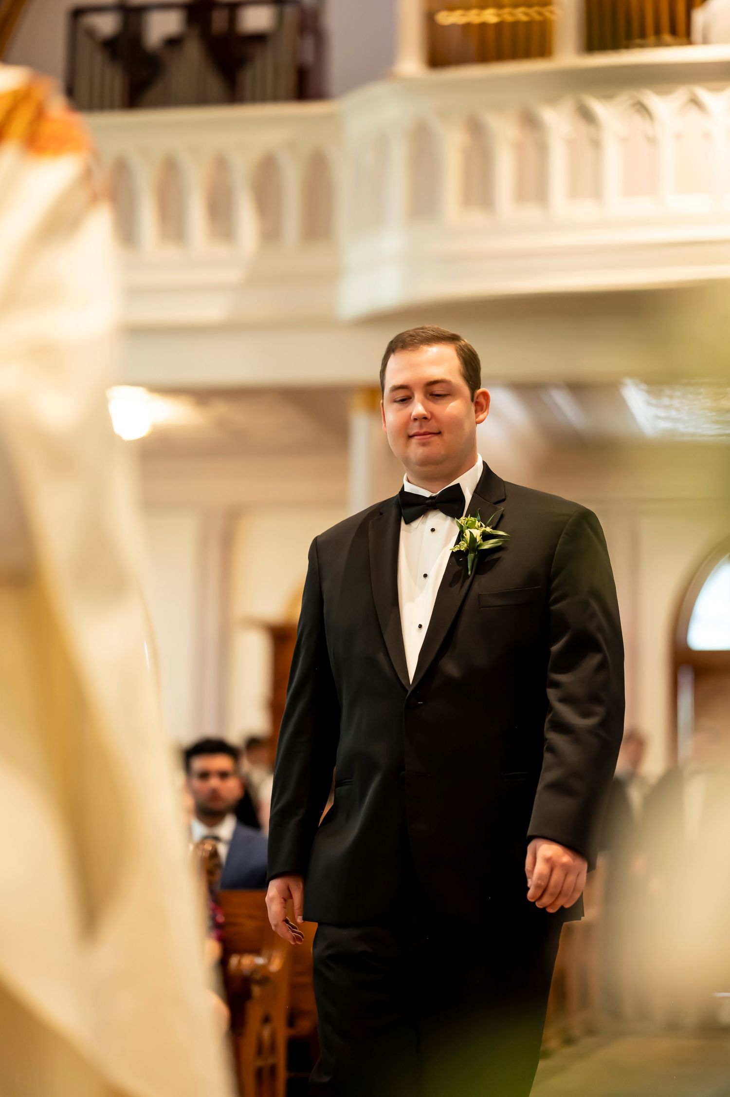 A groom in a classic black tuxedo with white boutonniere stands waiting at the altar in an elegant church setting.