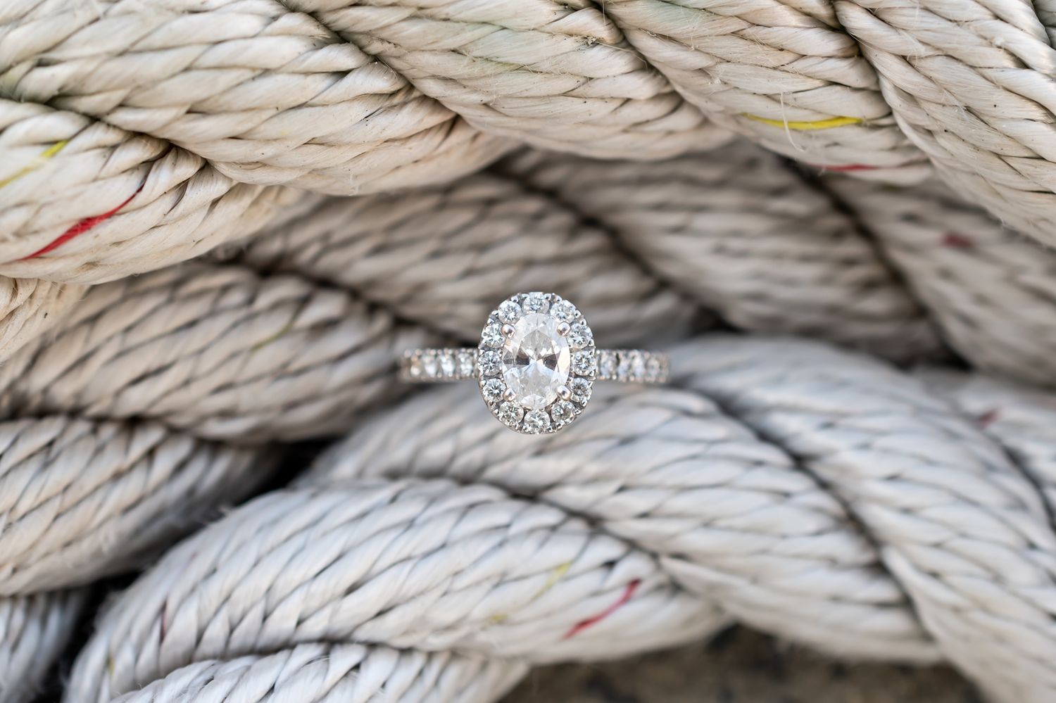 Sparkling oval engagement ring with diamond halo displayed against twisted beige nautical rope background.