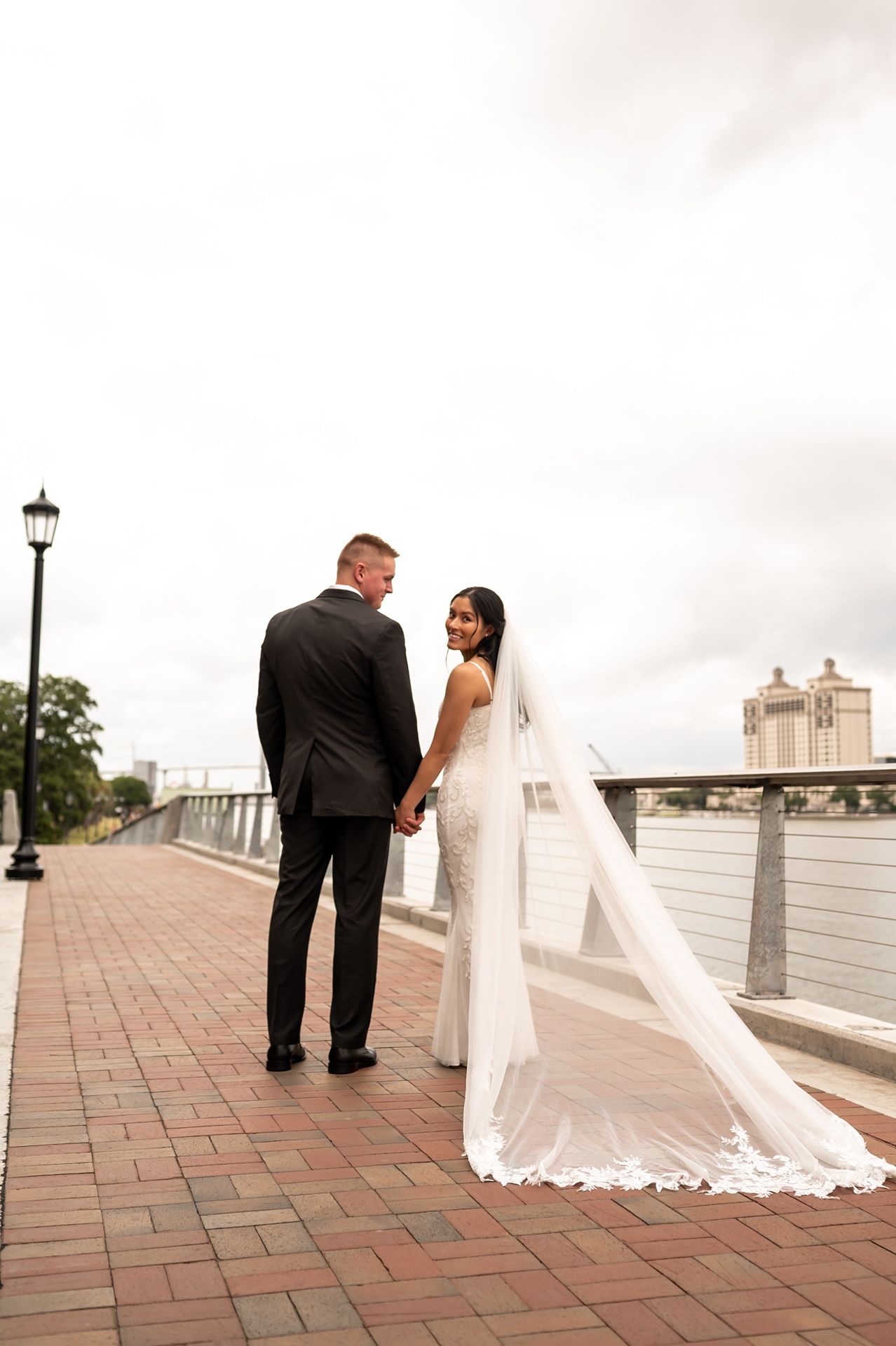 A couple shares a romantic moment on a brick walkway along a waterfront with a long flowing bridal veil.