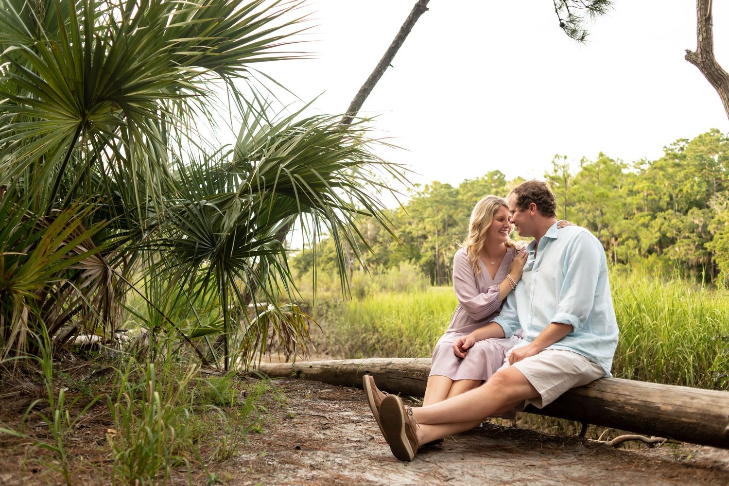 A couple shares an intimate moment while sitting together on a fallen log near palm trees and marsh grass.
