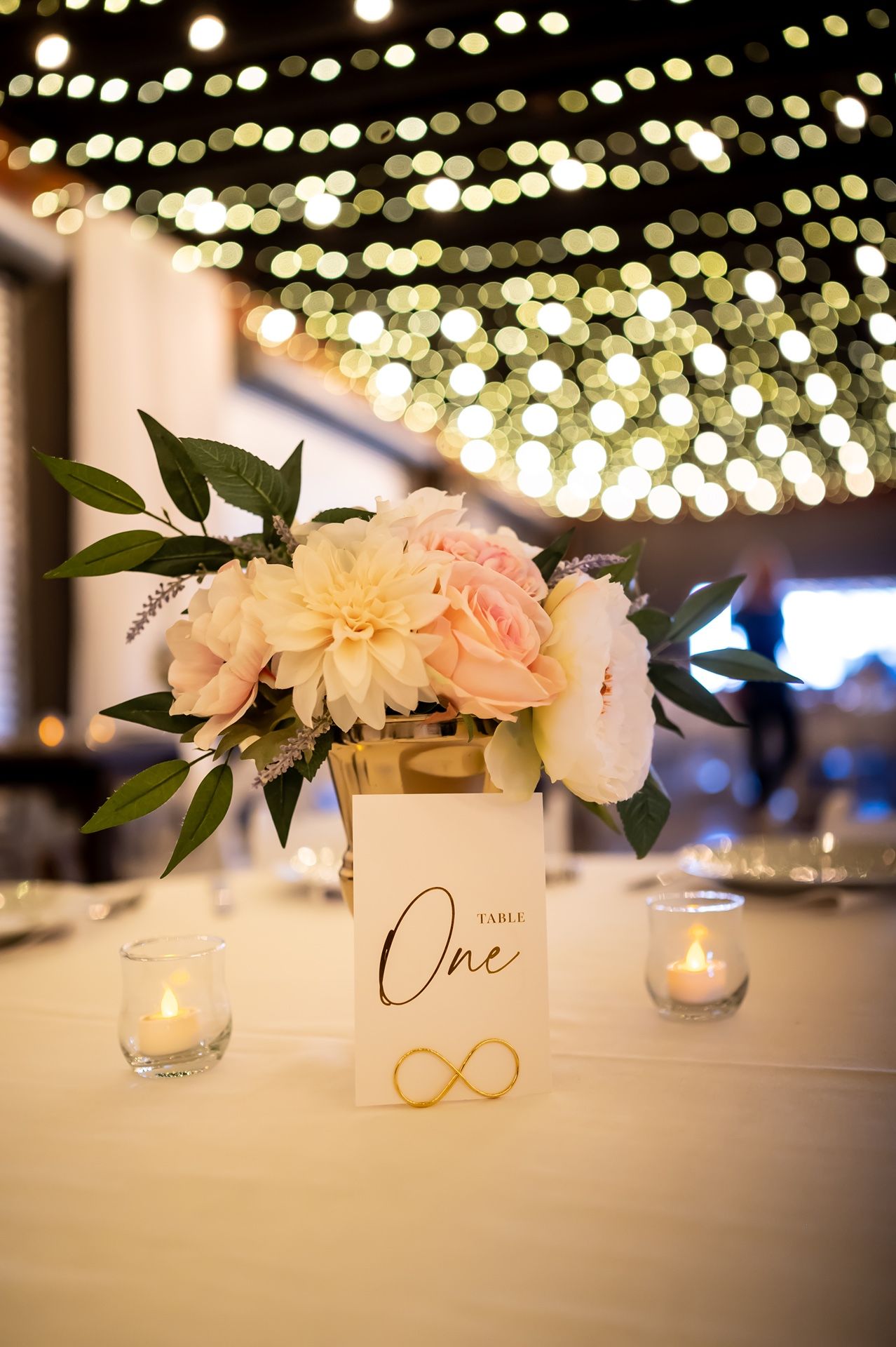 Wedding reception table details with soft string lights overhead and white floral centerpieces with gold accents and candles.