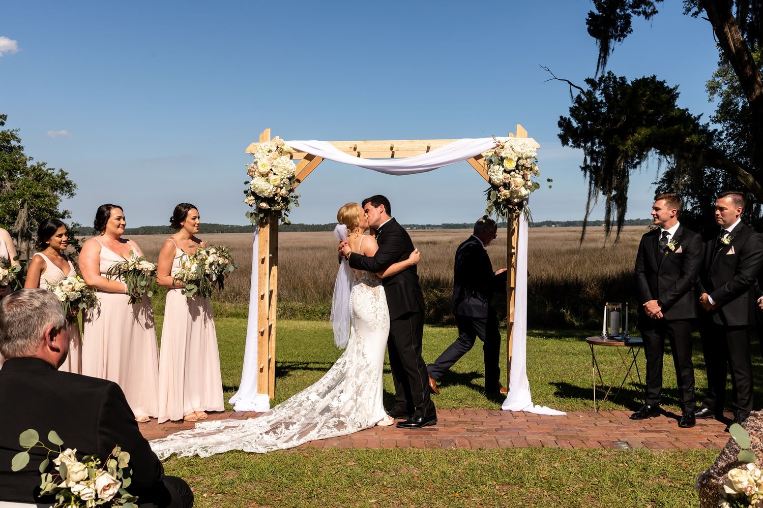 A panoramic wedding ceremony setup in a sunny outdoor field shows chairs, floral arrangements, and a decorative arch.
