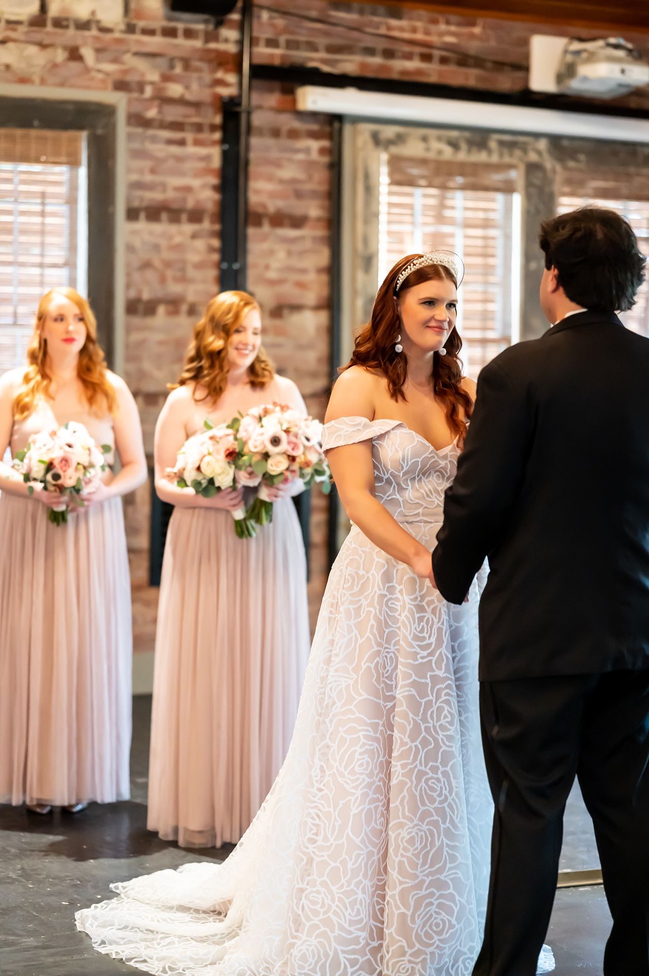 A bride and bridegroom exchange vows at a rustic brick venue while bridesmaids in blush gowns hold bouquets nearby.