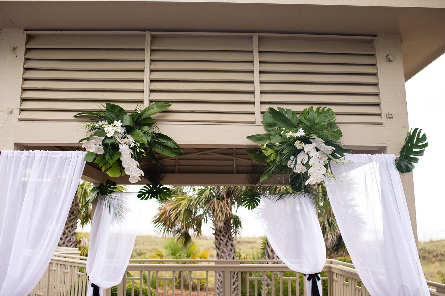 White sheer curtains and tropical greenery decorate an elegant outdoor wedding venue with palm trees in the background.