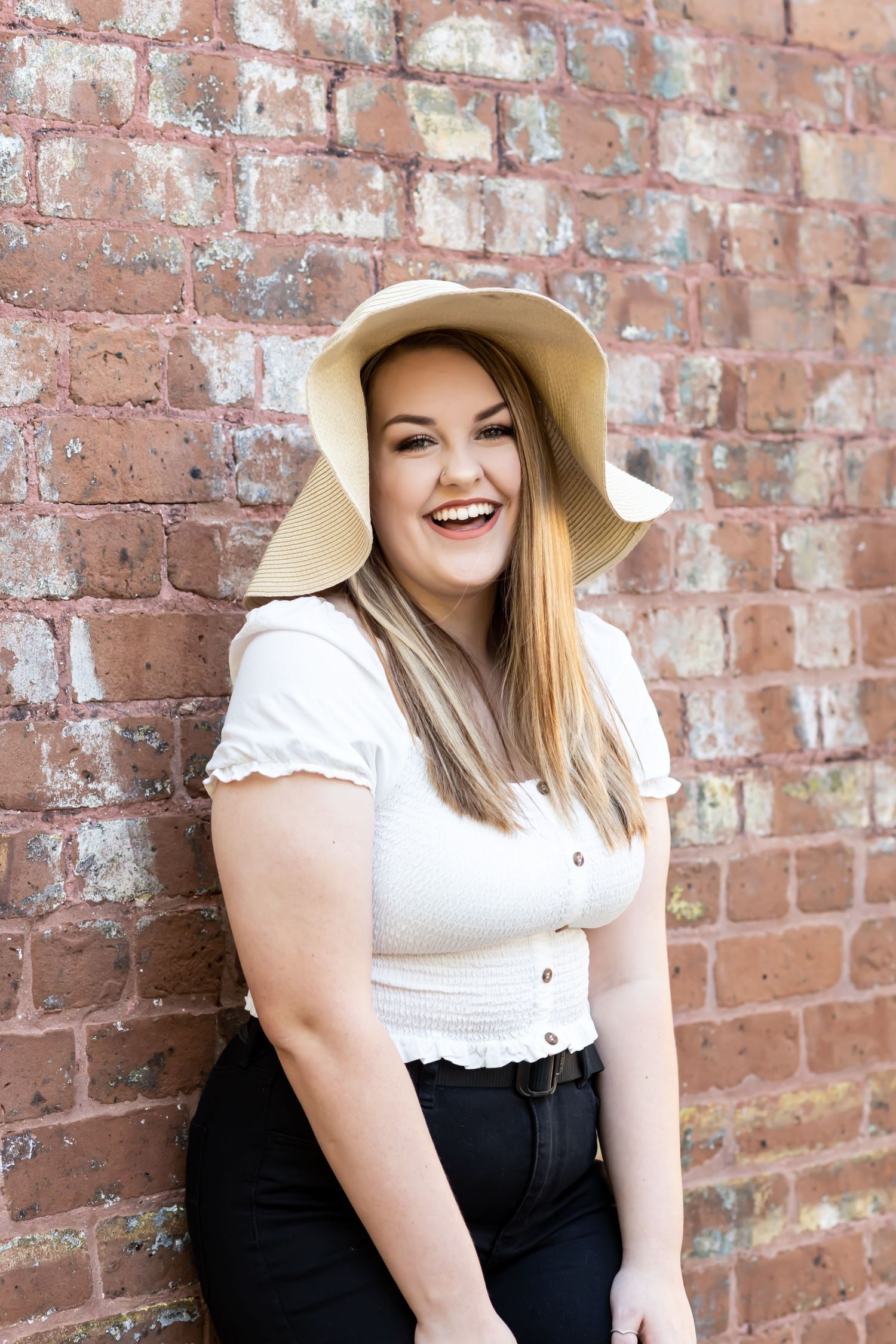 Stylish summer senior portrait with floppy hat against brick wall background.