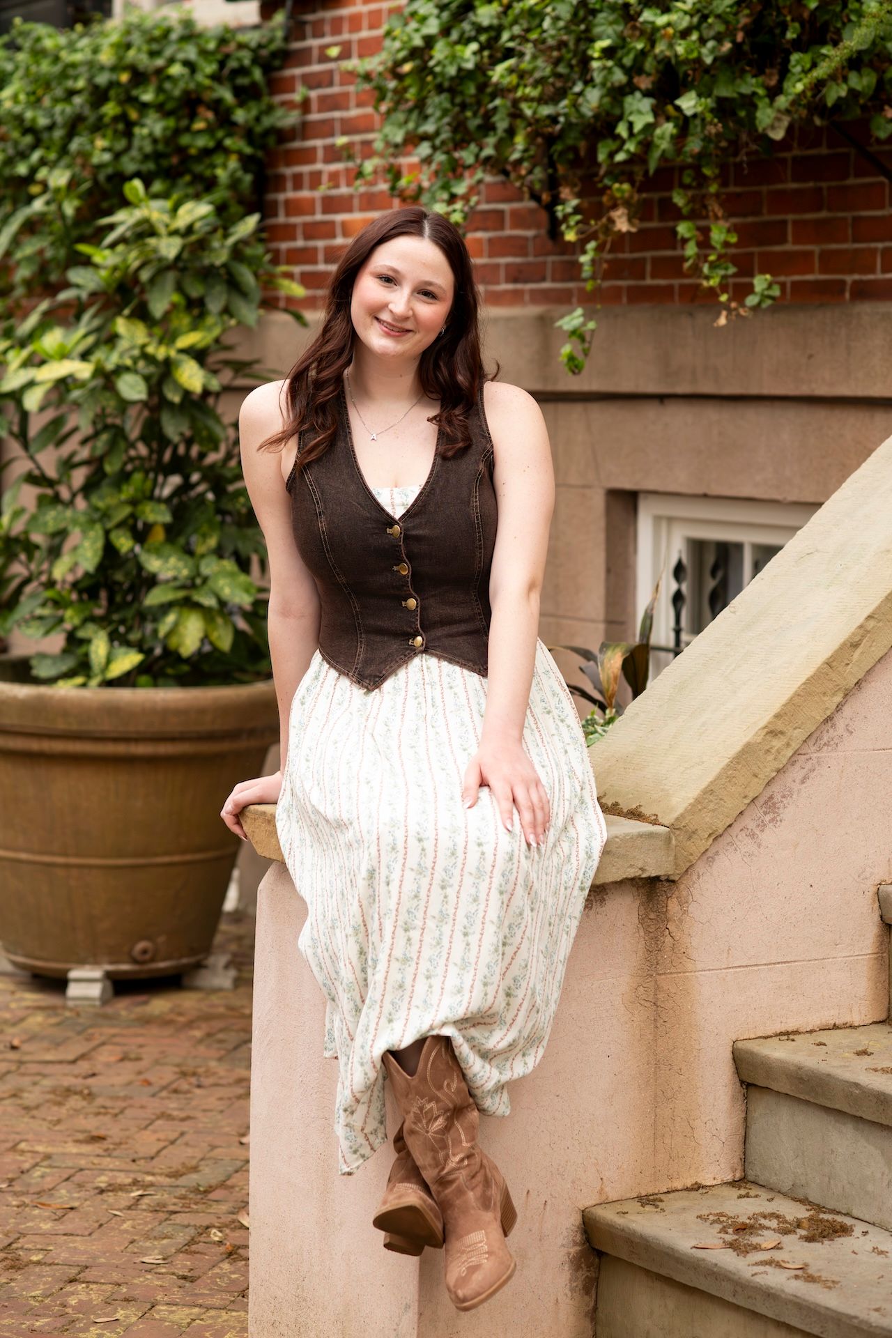 High school senior in brown vest, floral skirt, and cowboy boots sitting on stone steps near brick building with greenery on Jones Street.