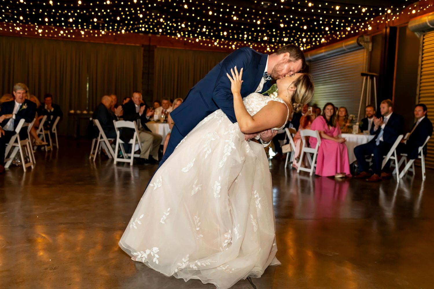 A couple share their first dance under sparkling string lights at their wedding reception with guests seated in the background.