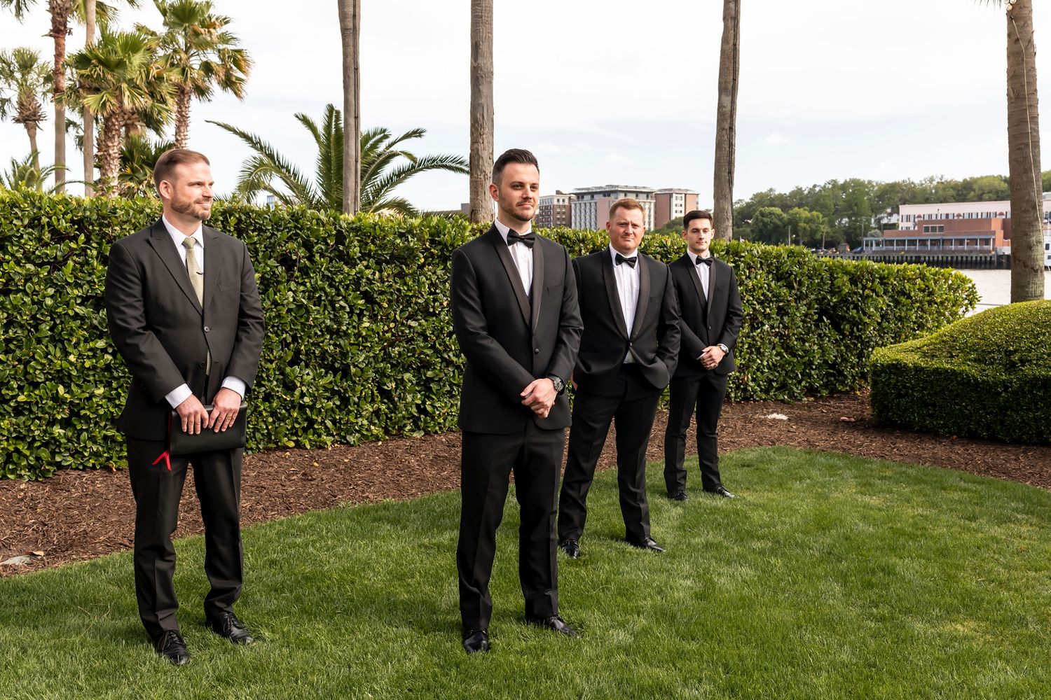Groomsmen in formal black tuxedos posed in front of lush green landscaping at an outdoor venue.