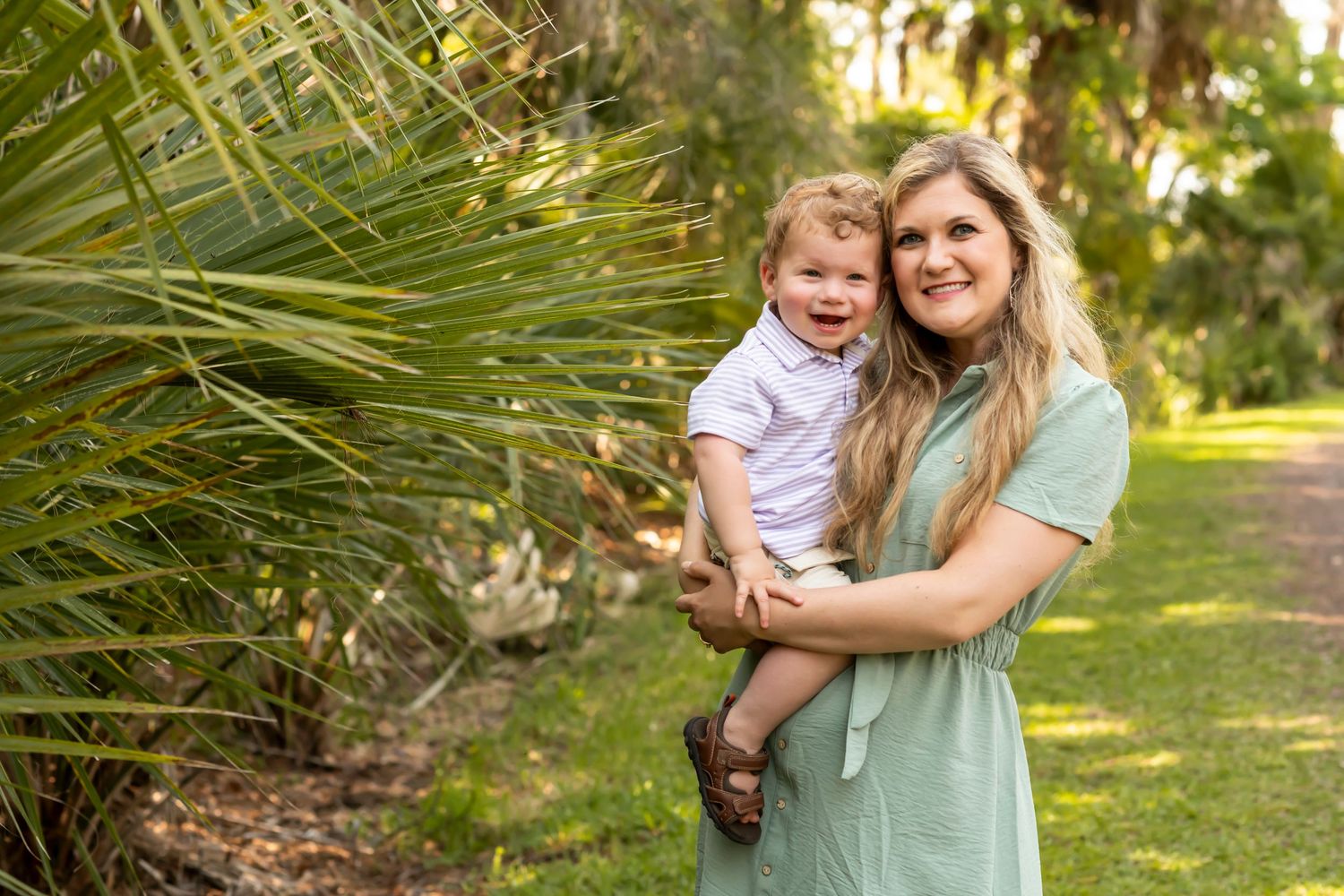 A parent in a sage green dress stands on a garden path near tropical plants and palm fronds.