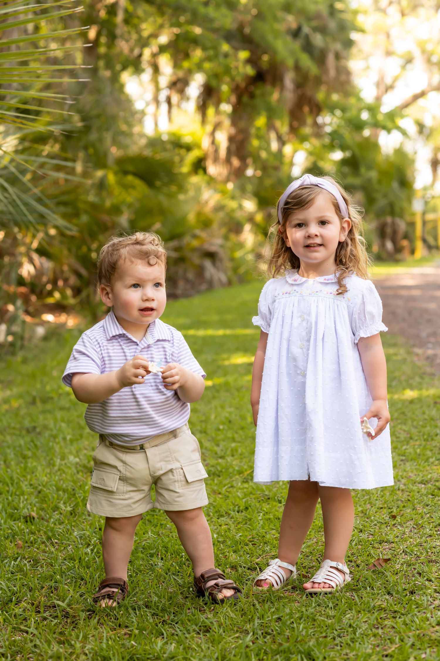 Two young children stand together on grass wearing summer outfits in a garden setting with trees in background.