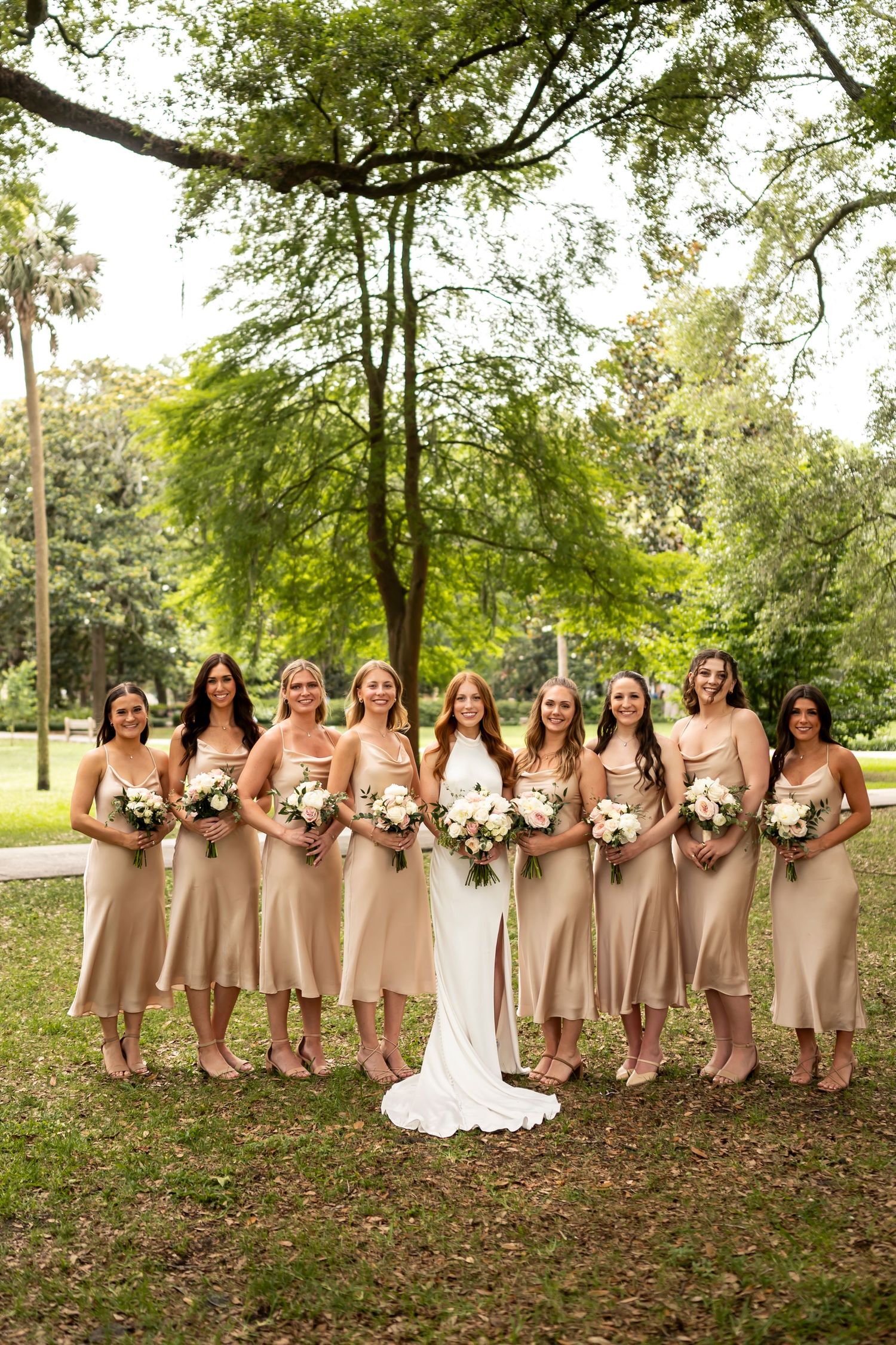 Group of bridesmaids in champagne silk dresses stand with bride holding bouquets in a garden setting under trees.
