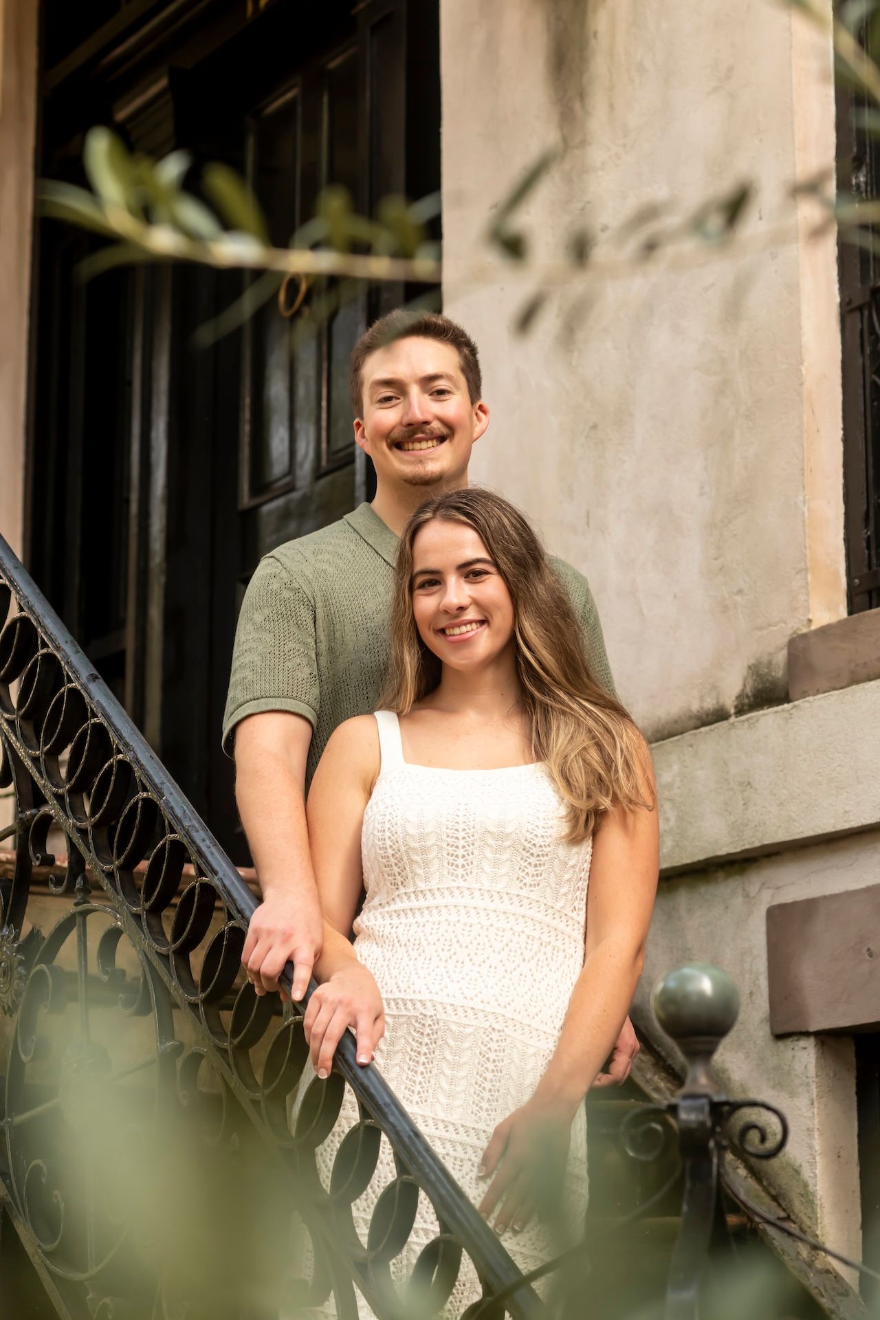 A couple stands together on outdoor stone steps with wrought iron railing, surrounded by soft greenery in natural light.