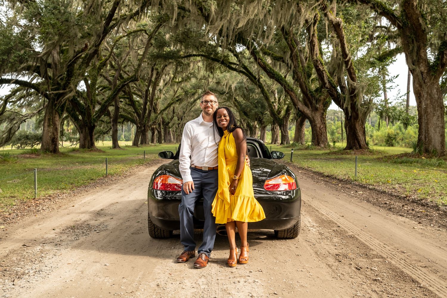 A couple stands in front of a luxury vehicle on a dirt road under a canopy of Spanish moss-draped oak trees.