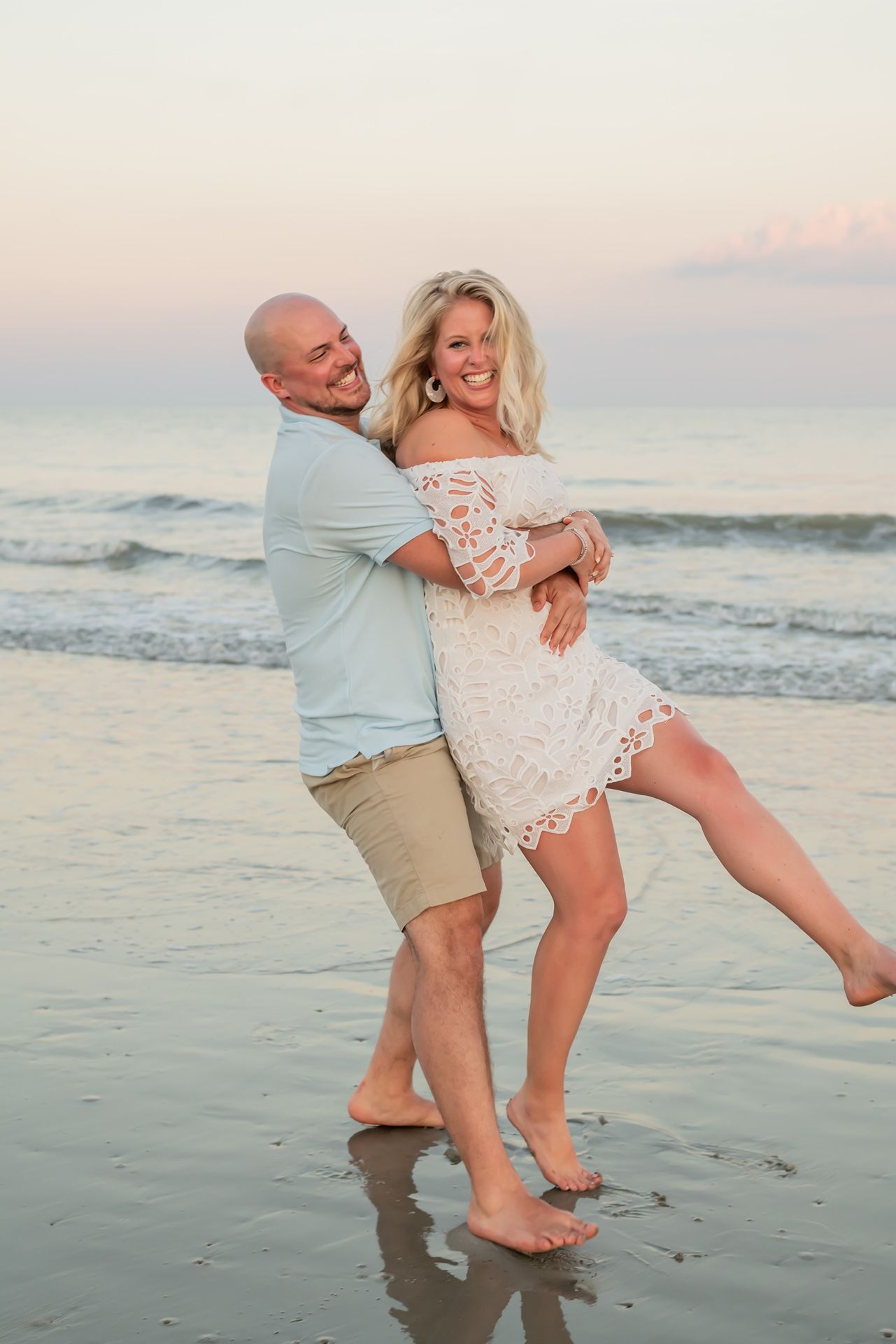 A couple in casual beach attire shares a playful moment while dancing on the shoreline at sunset.