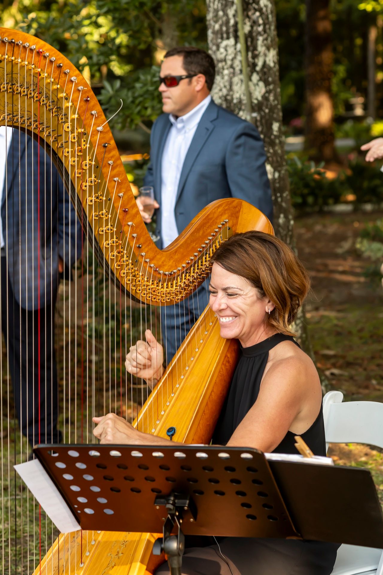 A musician playing a golden harp at an outdoor event with a music stand and trees in the background.