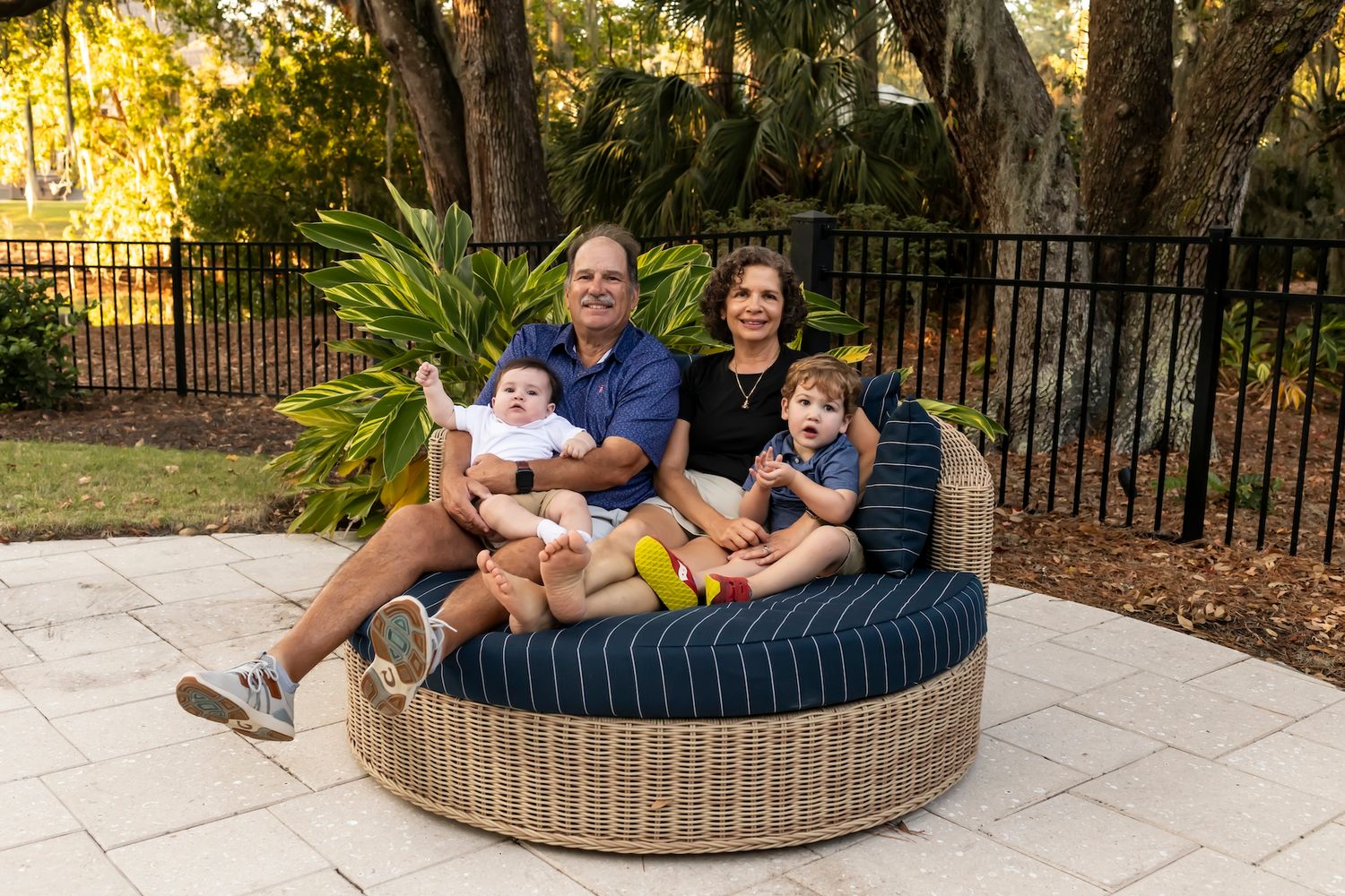 A group sits together on a round wicker patio daybed on a stone patio surrounded by iron fencing and lush greenery.