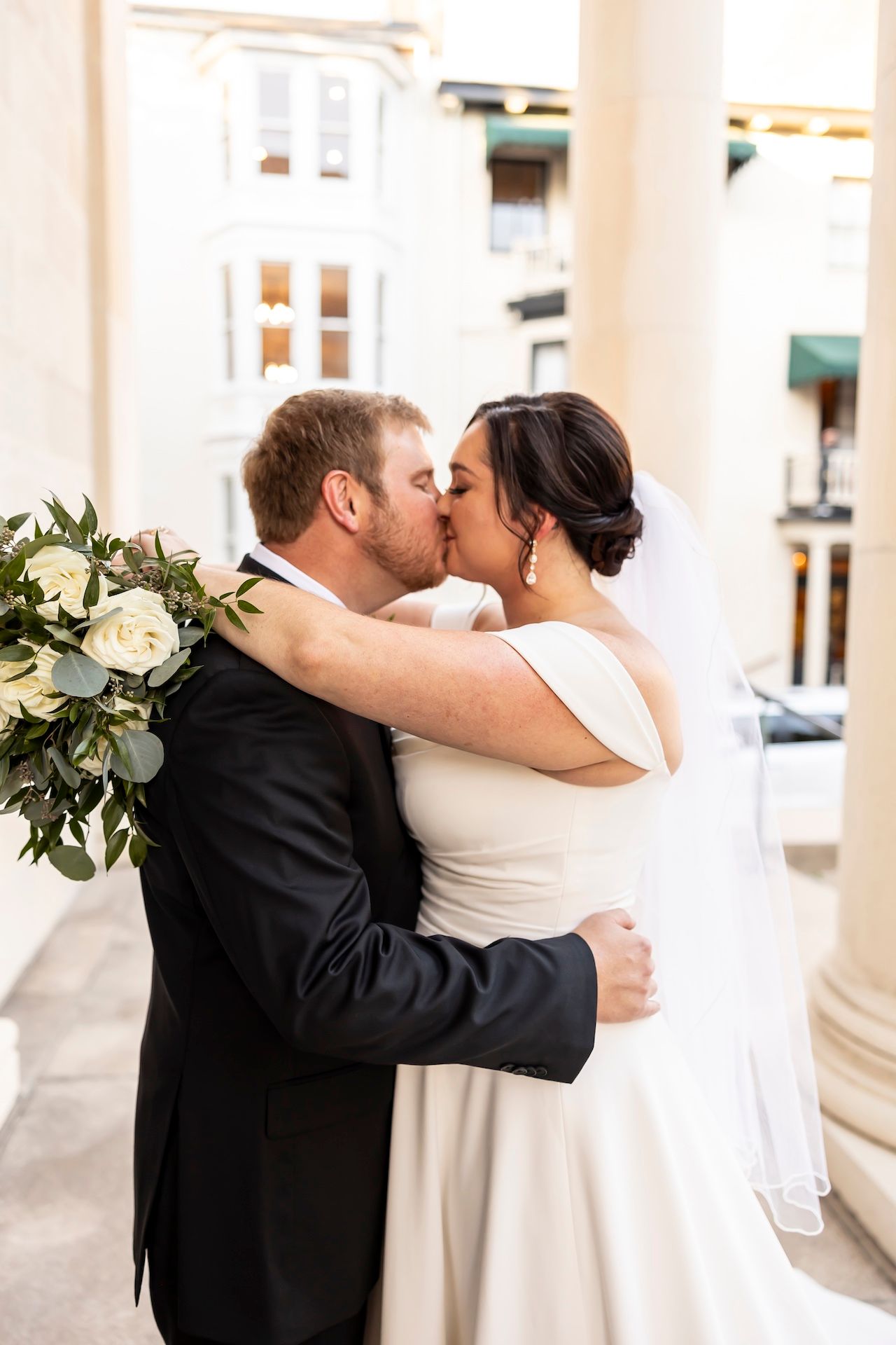 A newlywed couple in wedding attire share a kiss while embracing outdoors, with a white floral bouquet in view.