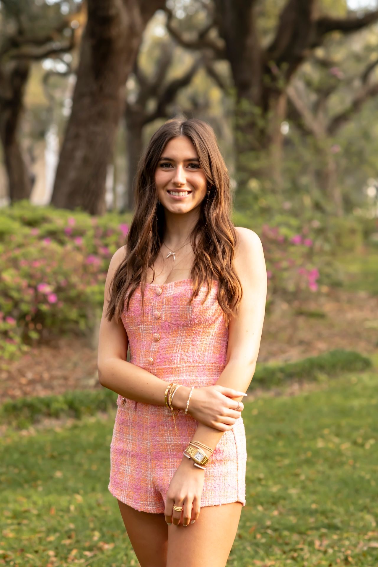 A person in a pink patterned romper poses outdoors in a garden setting with flowering trees and greenery.