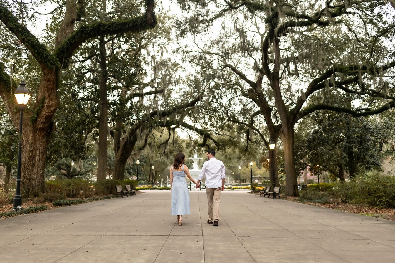 Two people in light colored clothing walk hand in hand down a scenic path lined with moss-draped oak trees at sunset.