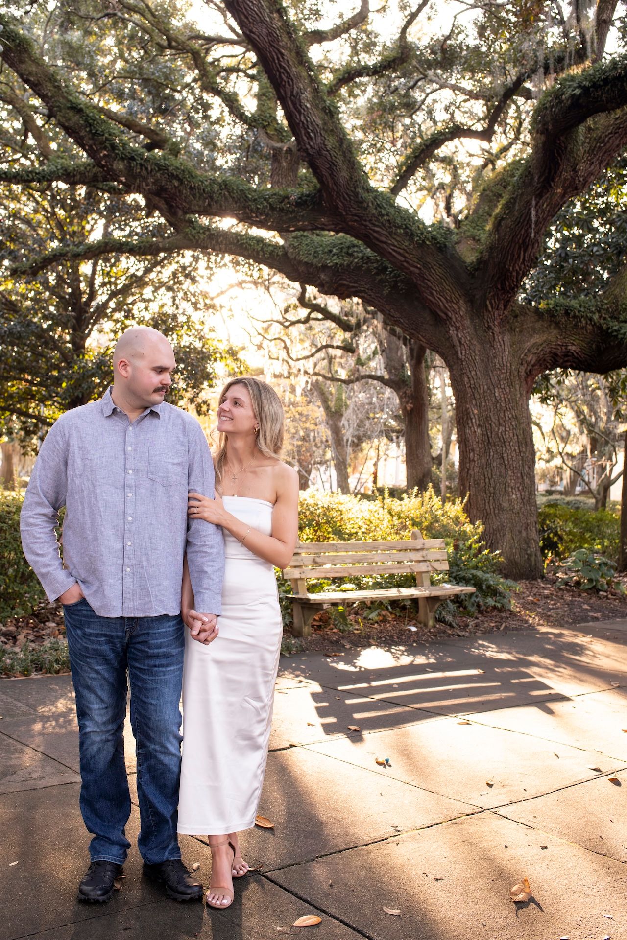 Two people holding hands while walking together under moss-draped oak trees in a romantic park setting at sunset.