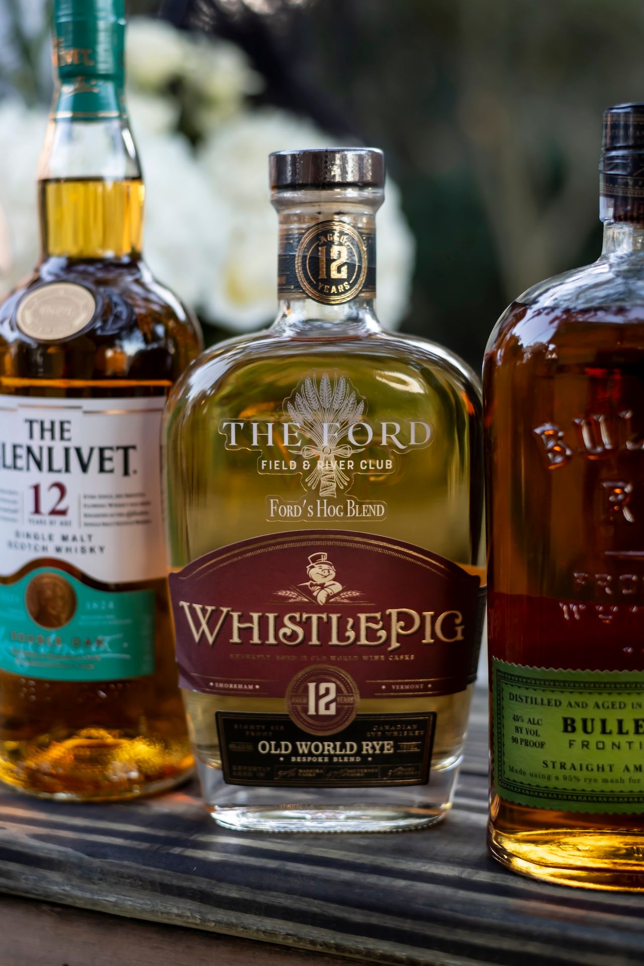 Three bottles of premium whiskey including The Glenlivet 12, WhistlePig Rye, and Bulleit displayed on a wooden surface.
