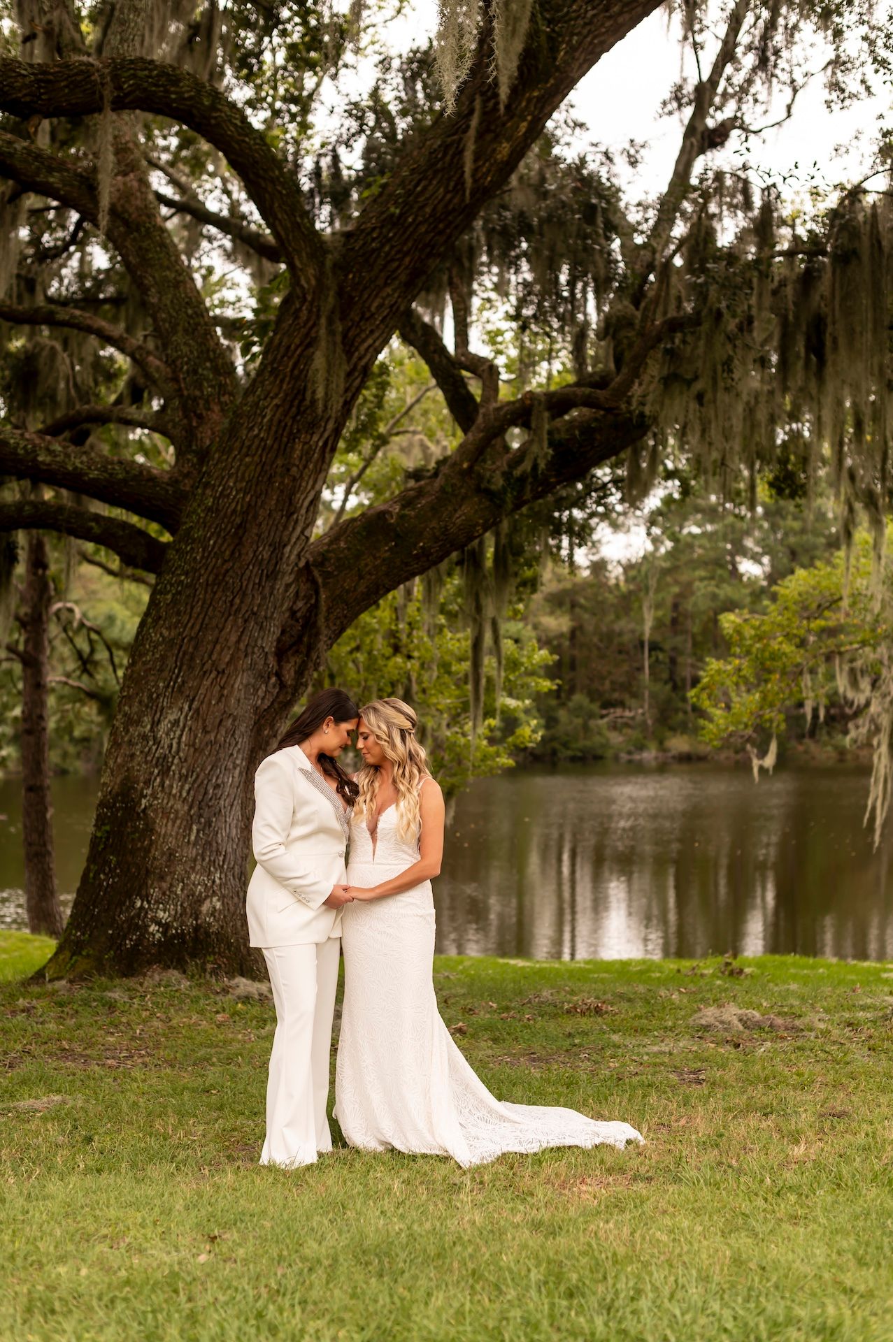 A romantic couple in white attire embrace under a majestic Spanish moss-draped oak tree beside a serene lake at sunset.