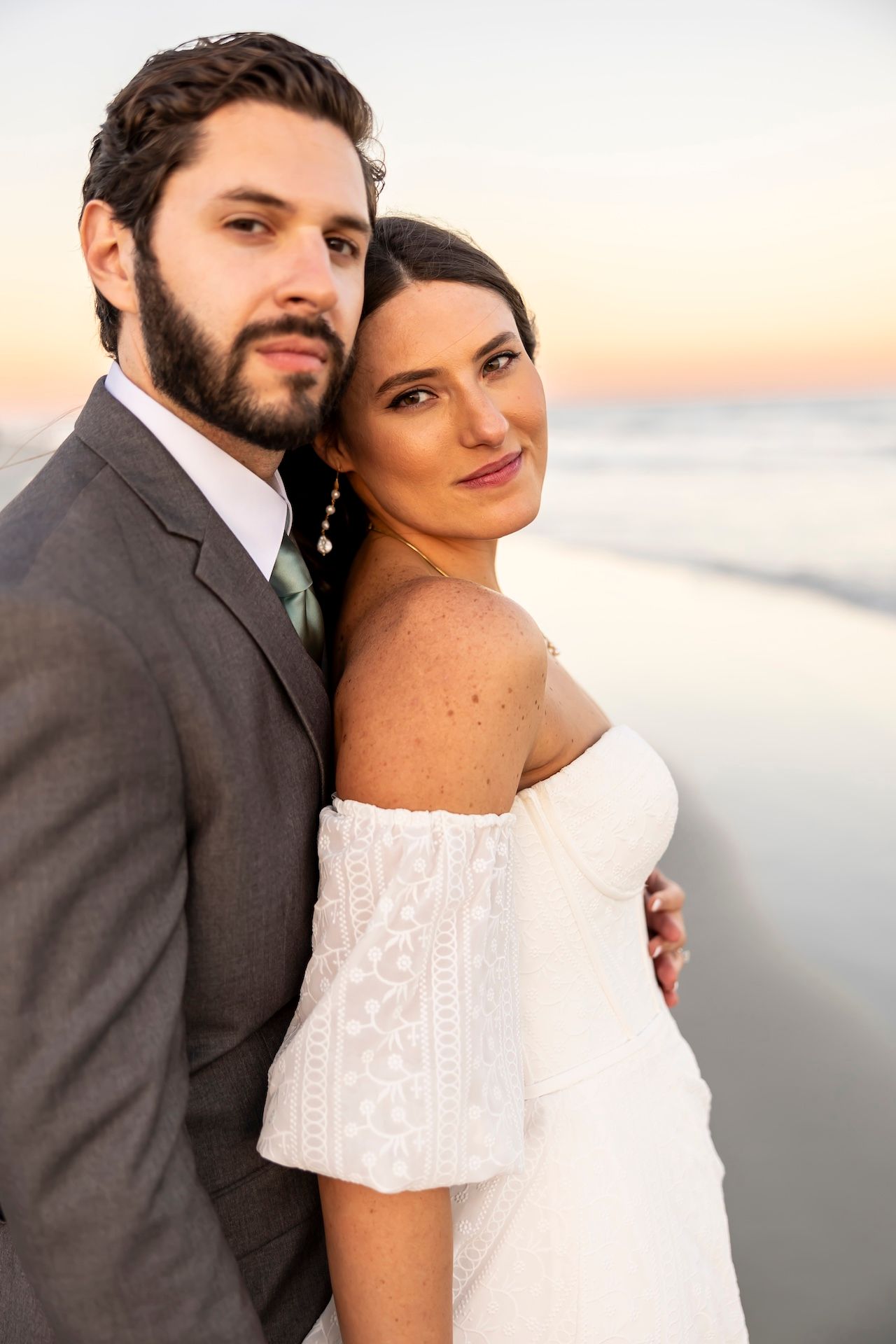 A couple poses at sunset by the beach, with the bride wearing an off-shoulder white dress and groom in a gray suit.